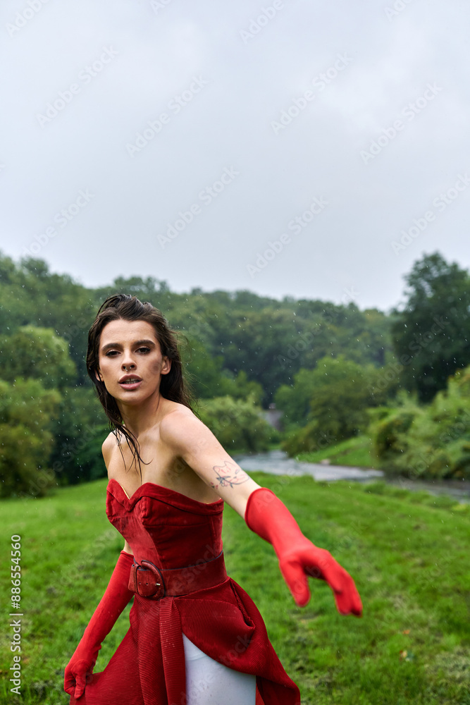 Obraz premium An attractive young woman in a flowing red dress and long gloves stands gracefully in a lush green field, enjoying the summer breeze.