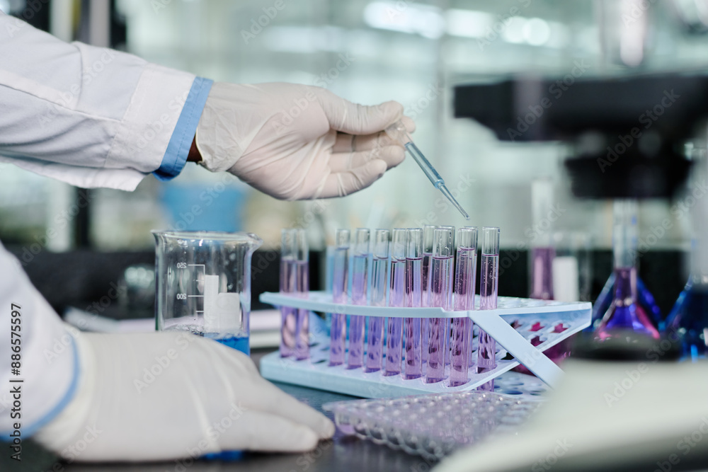Gloved hand of modern clinician standing by workplace in front of camera and dropping liquid substance into flasks from plastic pipette Gloved hand of modern clinician standing by workplace in front of camera and dropping liquid substance into flasks from plastic pipette