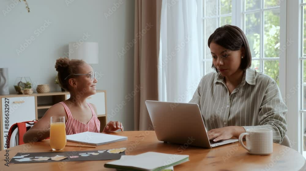 African American mother and daughter sitting at table at home. Black girl is drawing and puts on moms eyeglasses, woman is working on laptop and talks with kid smiling and enjoying African American mother and daughter sitting at table at home. Black girl is drawing and puts on moms eyeglasses, woman is working on laptop and talks with kid smiling and enjoying