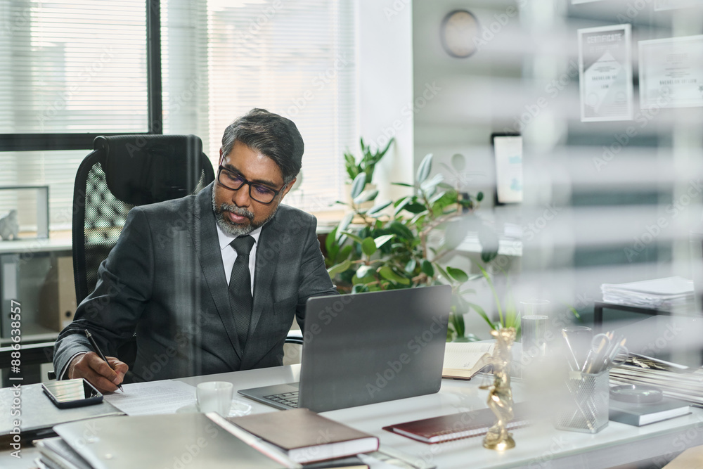 Obraz premium Serious mature male lawyer sitting by workplace in front of laptop, reading paper document and putting his signature below text