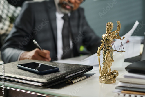 Statuette of Themis standing on workplace of lawyer with mobile phone on top of stack of folders containing juridical paper documents