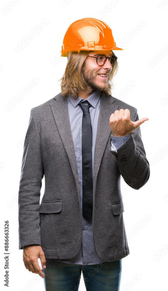 Young handsome architec man with long hair wearing safety helmet over isolated background smiling with happy face looking and pointing to the side with thumb up. Young handsome architec man with long hair wearing safety helmet over isolated background smiling with happy face looking and pointing to the side with thumb up.
