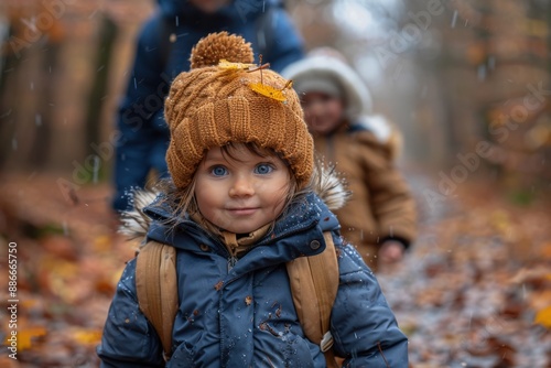 A young girl with piercing blue eyes smiles brightly as she walks on a snowy forest trail, adorned in winter gear and accompanied by family in a serene winter setting. photo