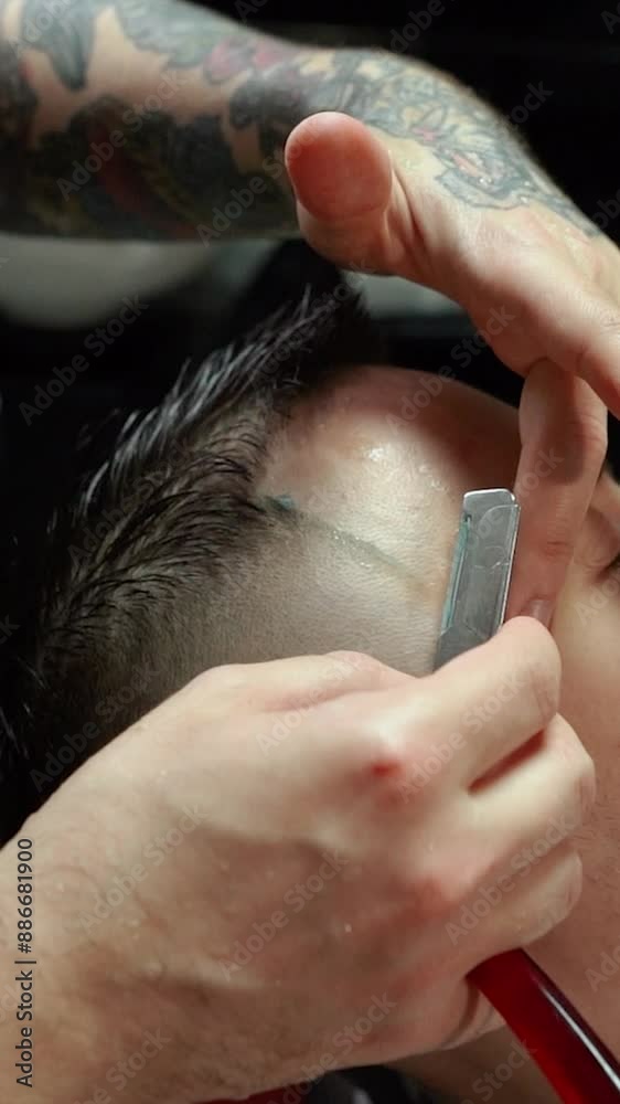 A barber man with a tattoo on his arm makes a haircut to a brunette guy in a barbershop, cuts his hair with a clipper close-up