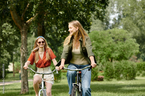 Obraz na plátně Two young women, dressed casually, ride their bikes through a lush green park on a sunny day, smiling and enjoying each other company