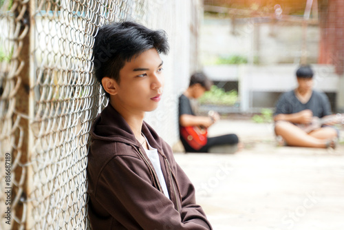 Asian male teenager sits with his back against a metal fence wall in a juvenile detention center. Concept: detention, freedom, imprisonment, confinement, reformation, loneliness.