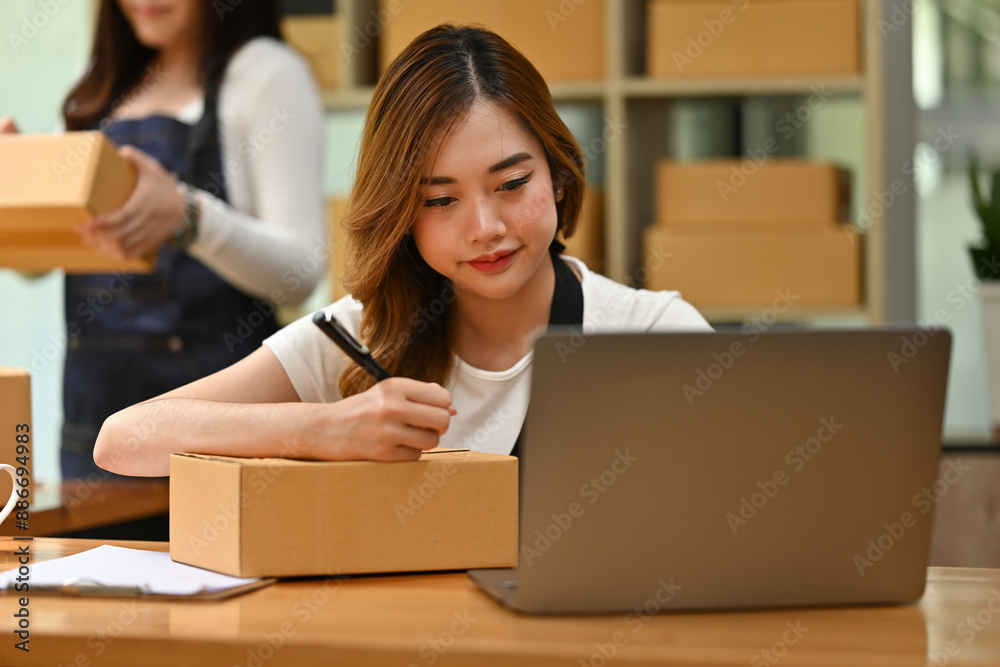 A woman uses a laptop to manage online orders while writing an address on a package, with another person in the background handling more boxes, integrating digital and manual processes in SME A woman uses a laptop to manage online orders while writing an address on a package, with another person in the background handling more boxes, integrating digital and manual processes in SME