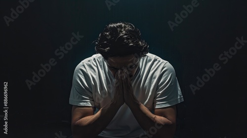A man with his head bowed in a dark room, wearing a white t-shirt with both hands on his knees praying against a black background.