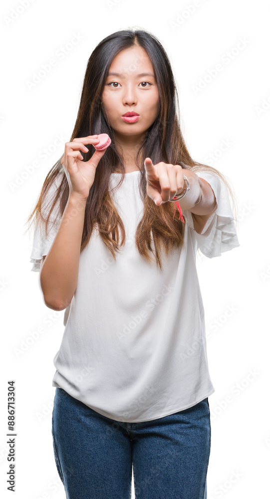 Young asian woman eating pink macaron sweet over isolated background pointing with finger to the camera and to you, hand sign, positive and confident gesture from the front Young asian woman eating pink macaron sweet over isolated background pointing with finger to the camera and to you, hand sign, positive and confident gesture from the front