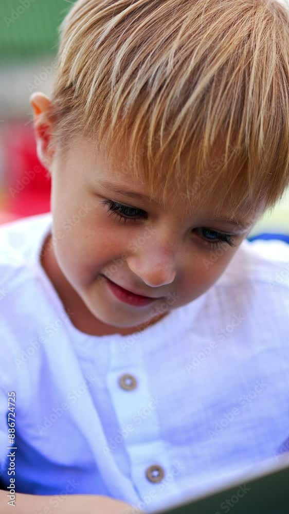 Lovely fair-haired little boy spending time outdoors. Caucasian kid sitting calmly in chair with laptop, pressing keys on keyboard. Close up. Vertical video Lovely fair-haired little boy spending time outdoors. Caucasian kid sitting calmly in chair with laptop, pressing keys on keyboard. Close up. Vertical video