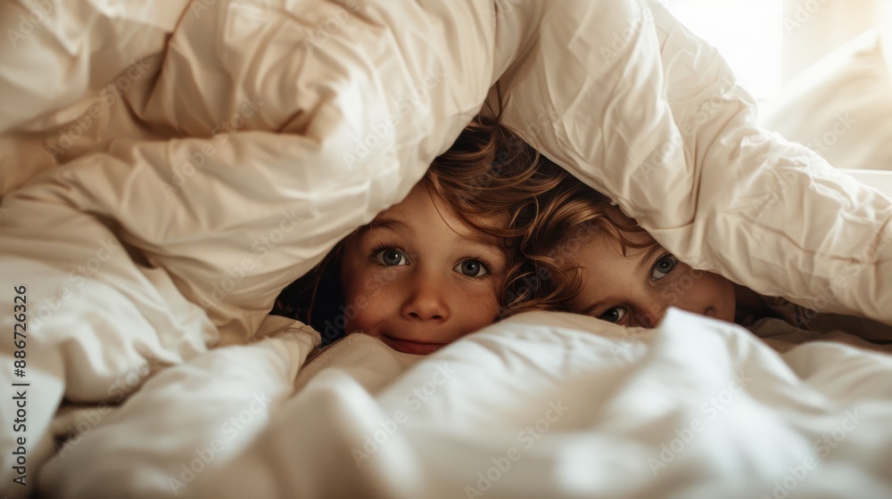 Two young children with curly hair are peeking out from under a fluffy white blanket, creating a heartwarming and cozy moment of childhood cheer and warmth. Two young children with curly hair are peeking out from under a fluffy white blanket, creating a heartwarming and cozy moment of childhood cheer and warmth.