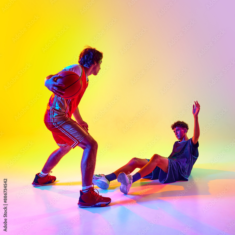 Two basketball players in action, with one player dribbling and other fallen on floor, in neon light against gradient studio background. Concept of professional sport, competition, match, team games. Two basketball players in action, with one player dribbling and other fallen on floor, in neon light against gradient studio background. Concept of professional sport, competition, match, team games.