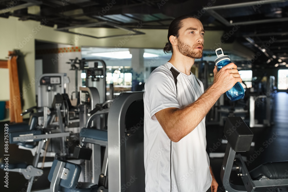 An athletic man in activewear takes a break to drink water from a bottle while working out in the gym. An athletic man in activewear takes a break to drink water from a bottle while working out in the gym.