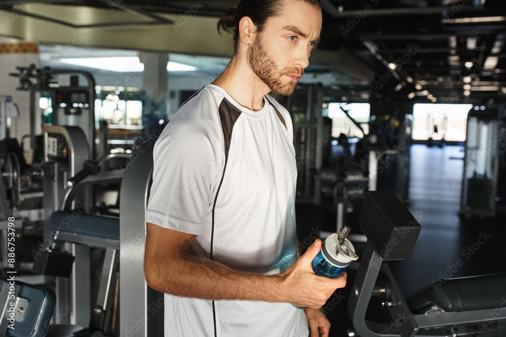 An athletic man in active wear takes a break, holding a bottle of water in a gym surrounded by exercise equipment.