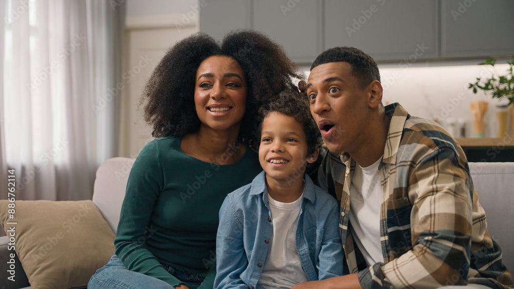 Obraz premium Excited happy playful African American ethnic family mother father embracing little boy child playing game parents kid watching funny TV show movie posing making faces grimacing indoors home apartment