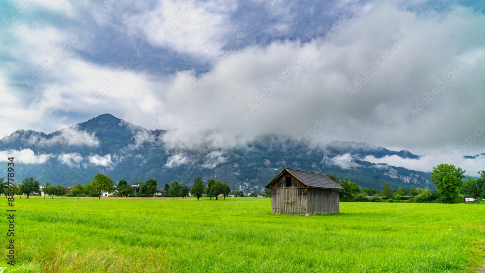After a thunderstorm, scenery with barn and cabin on green field and sun rays through cloud, light and dark green grass after a long storm, white clouds under dark Thunderstorm After a thunderstorm, scenery with barn and cabin on green field and sun rays through cloud, light and dark green grass after a long storm, white clouds under dark Thunderstorm