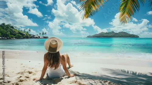 A woman in a white dress and sunhat enjoys a relaxing day on a tropical beach, sitting by the turquoise waters and under a bright blue sky. The image evokes a sense of peace and a perfect beach getawa photo