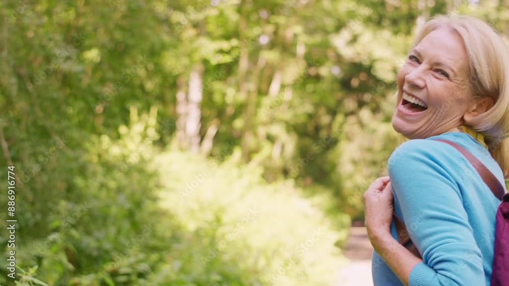 Rear view of mature woman wearing backpack walking ahead of camera along path through trees and looking back over shoulder - shot in slow motion Rear view of mature woman wearing backpack walking ahead of camera along path through trees and looking back over shoulder - shot in slow motion