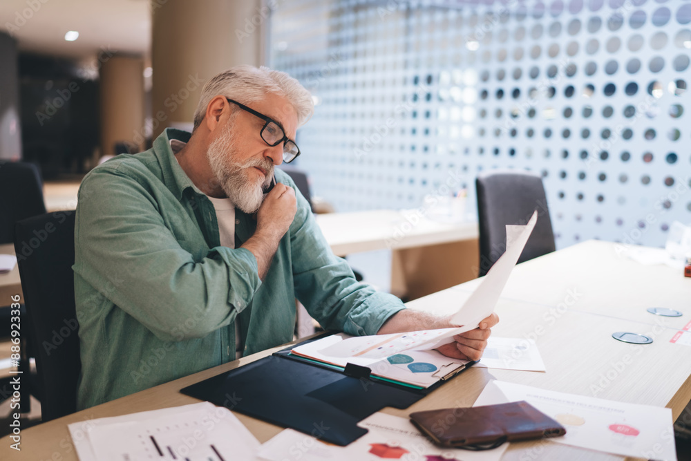 Older Caucasian man in a modern office setting, seated at desk and deeply engaged in analyzing business reports. Wearing glasses, focused on financial charts and graphs, professional environment Older Caucasian man in a modern office setting, seated at desk and deeply engaged in analyzing business reports. Wearing glasses, focused on financial charts and graphs, professional environment