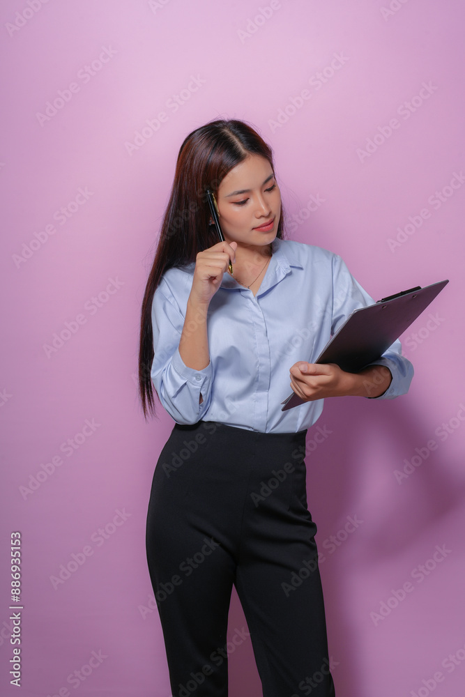 Focused young businesswoman reads a document, holding a clipboard and pen, showing determination and confidence against a pink background