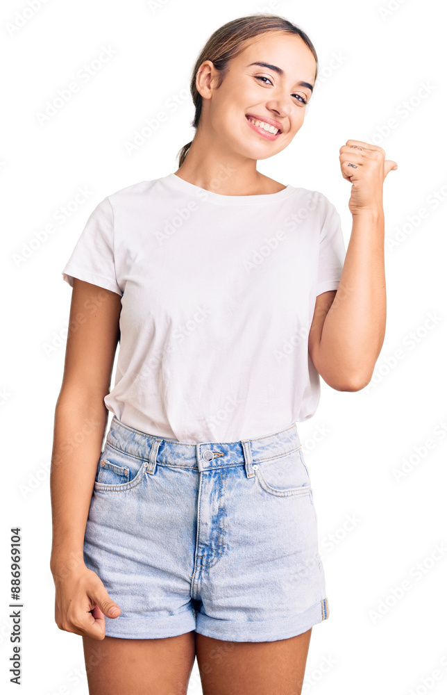 Young beautiful blonde woman wearing casual white tshirt smiling with happy face looking and pointing to the side with thumb up. Young beautiful blonde woman wearing casual white tshirt smiling with happy face looking and pointing to the side with thumb up.