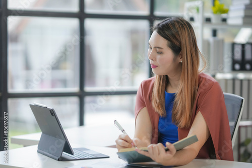 Focused asian businesswoman in modern office, taking notes on tablet. Natural light adds professional vibe. Productive and confident, showing ambition and motivation in remote work