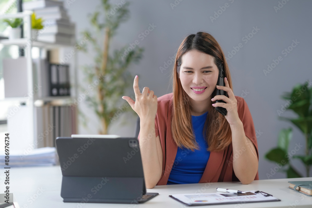 Cheerful asian businesswoman multitasks happily at her tidy desk in a bright office, talking on the phone and managing work tasks on her laptop confidently Cheerful asian businesswoman multitasks happily at her tidy desk in a bright office, talking on the phone and managing work tasks on her laptop confidently