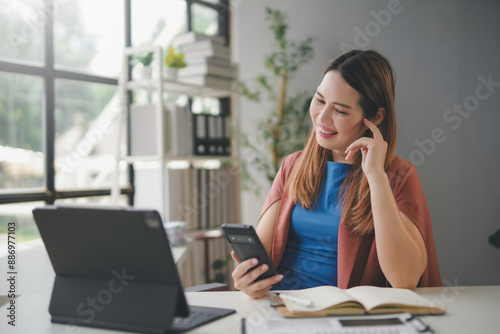 Young asian businesswoman multitasks happily at her modern office desk, using a smartphone and tablet. She looks professional and focused, surrounded by paperwork