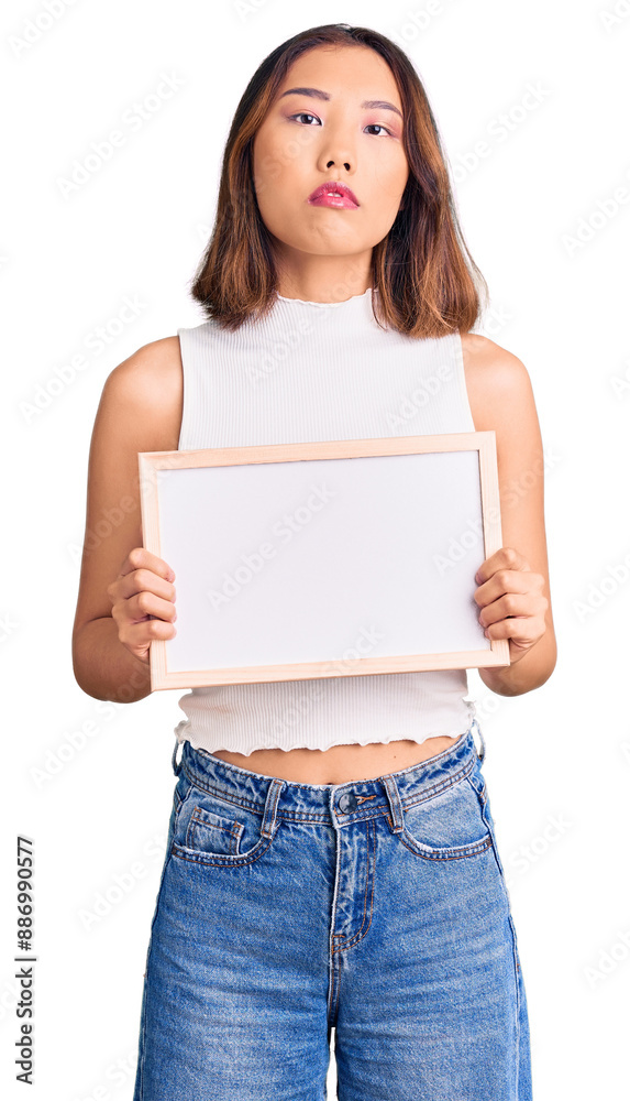 Young beautiful chinese girl holding empty white chalkboard thinking attitude and sober expression looking self confident