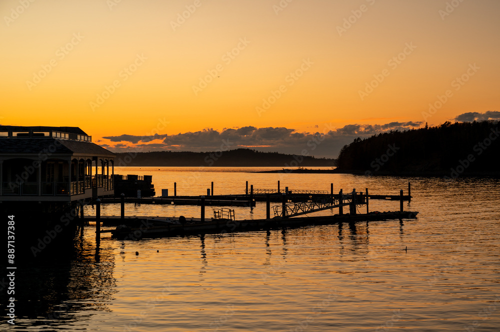 Sunset Over a Dock in Bar Harbor Maine. A calm, golden sunset casts long shadows across a wooden dock in Maine. The water is still and reflects the warm colors of the sky. A small building is silhoue Sunset Over a Dock in Bar Harbor Maine. A calm, golden sunset casts long shadows across a wooden dock in Maine. The water is still and reflects the warm colors of the sky. A small building is silhoue