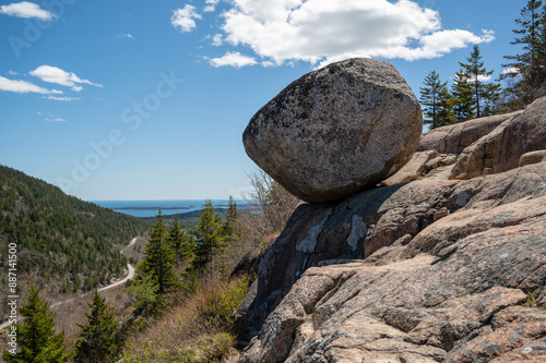 Precariously Balanced Bubble Boulder on a Mountainside Overlooking the Ocean. A large, round boulder rests precariously on a smaller rock formation, offering a breathtaking view of a winding road lea