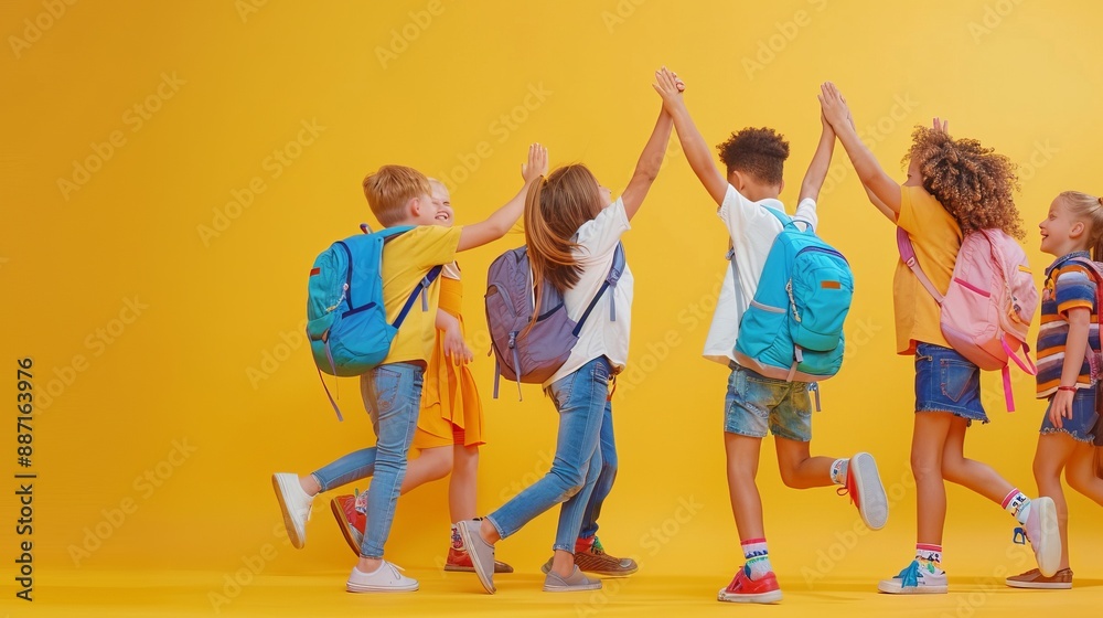 Fototapeta premium A joyful group of primary school kids wearing casual clothes and backpacks high-five each other in front of a bright yellow background, symbolizing unity and celebration.