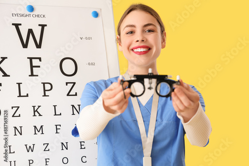 Female ophthalmologist with trial frame and eye test chart on yellow background, closeup photo