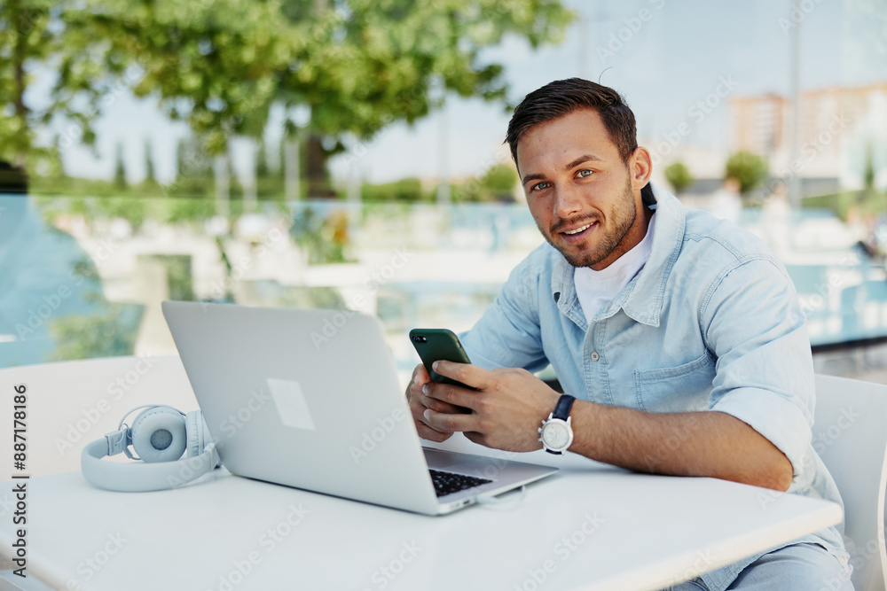Young man working remotely on laptop, smiling while using smartphone at outdoor cafe Young man working remotely on laptop, smiling while using smartphone at outdoor cafe