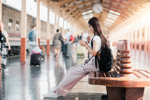 Wallpaper Mural Young woman sitting on a bench at a train station, using her smartphone. Travelers and luggage in the background, under a bright, sunlit roof. Torontodigital.ca