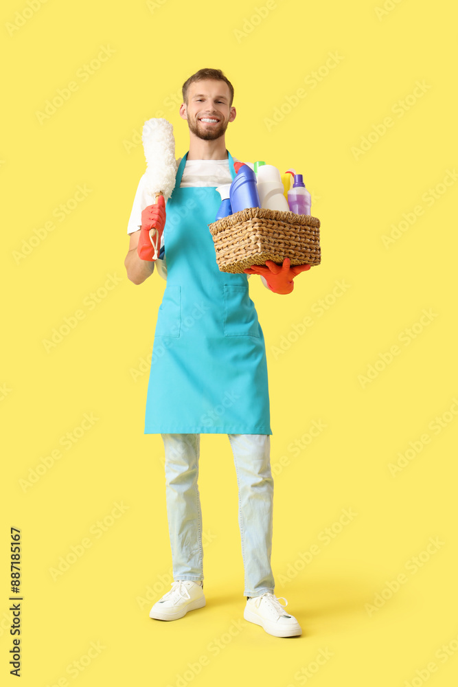 Male janitor holding dust pan and basket with cleaning supplies on yellow background Male janitor holding dust pan and basket with cleaning supplies on yellow background