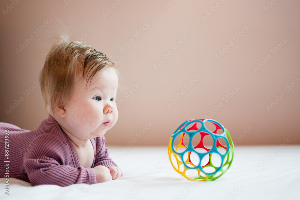 Cute Caucasian newborn baby girl during tummy time looking at the colorful ball. A 3-month-old girl with hair. Child care concept with copy space Cute Caucasian newborn baby girl during tummy time looking at the colorful ball. A 3-month-old girl with hair. Child care concept with copy space
