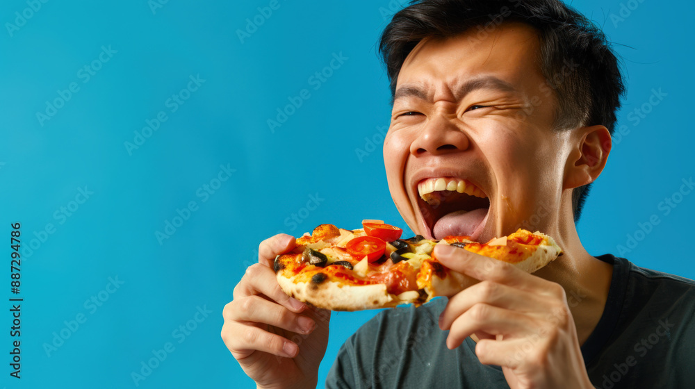 An Asian hungry man pounced on a piece of pizza with his mouth wide open. The pizza is decorated with tomatoes and olives. The guy is standing on a blue background An Asian hungry man pounced on a piece of pizza with his mouth wide open. The pizza is decorated with tomatoes and olives. The guy is standing on a blue background