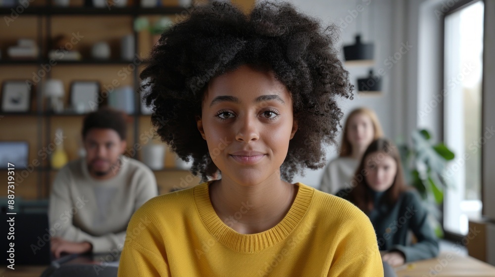 In a modern office, a young mixed-race woman with an afro and yellow sweater gazes calmly at the camera amidst a bustling background of coworkers In a modern office, a young mixed-race woman with an afro and yellow sweater gazes calmly at the camera amidst a bustling background of coworkers