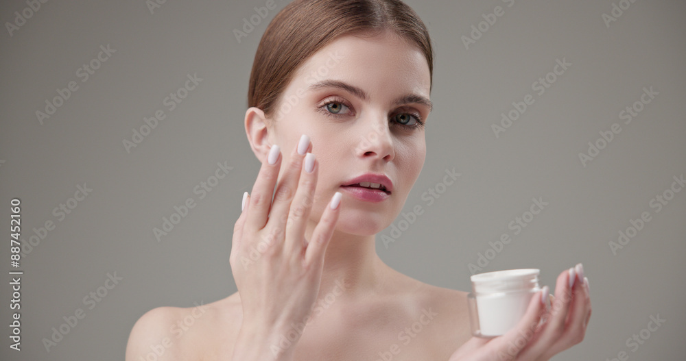 Beauty, health, cosmetics, anti-aging therapy and skin care concept - young beautiful brunette Caucasian woman holds a jar of moisturizer in her hands and applies the cream to her face skin Beauty, health, cosmetics, anti-aging therapy and skin care concept - young beautiful brunette Caucasian woman holds a jar of moisturizer in her hands and applies the cream to her face skin