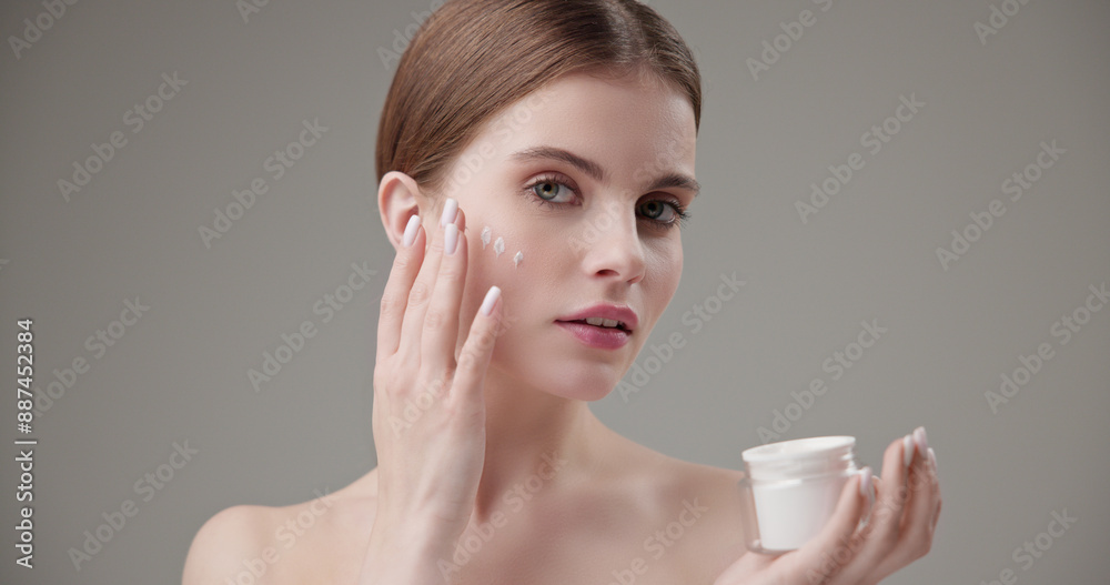Beauty, health, cosmetics, anti-aging therapy and skin care concept - young beautiful brunette Caucasian woman holds a jar of moisturizer in her hands and applies the cream to her face skin Beauty, health, cosmetics, anti-aging therapy and skin care concept - young beautiful brunette Caucasian woman holds a jar of moisturizer in her hands and applies the cream to her face skin