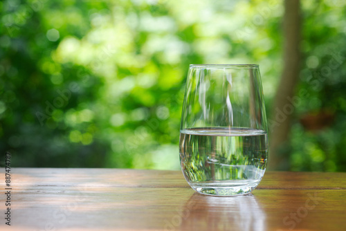 half glass of drinking water on wooden table in garden with green bokeh background.