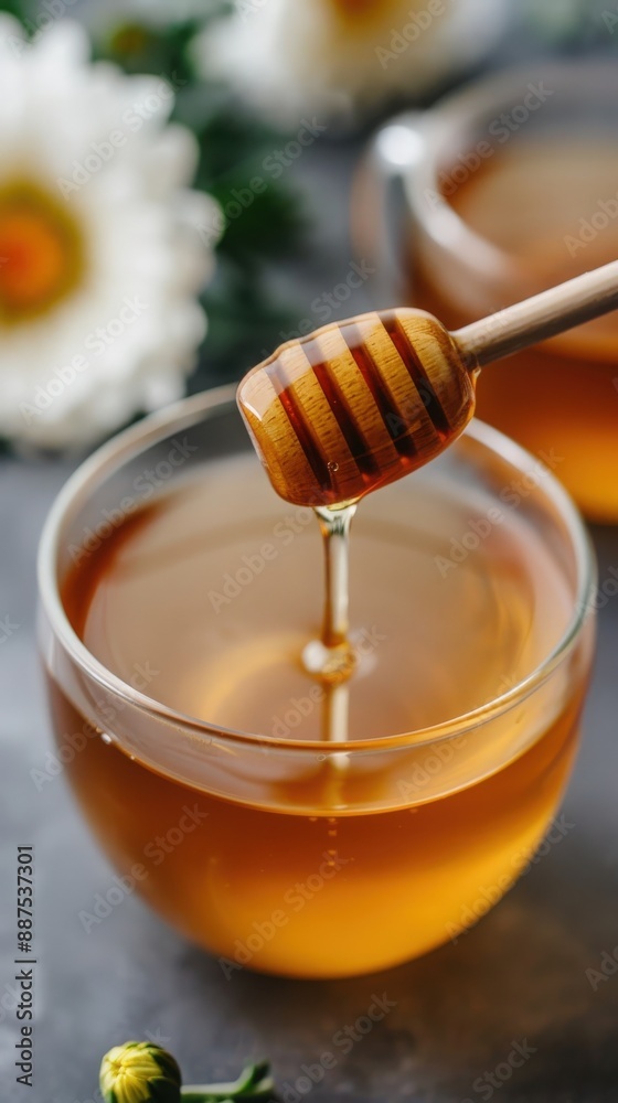 Close-up of golden honey dripping into a clear glass cup, with a flower and honey dipper in the background. Close-up of golden honey dripping into a clear glass cup, with a flower and honey dipper in the background.