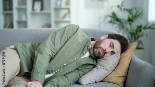 Exhausted man tired after hard working day lies down on sofa in living room at home. Overworked overdriven male wants to sleep, falls off his feet on couch, puts his head on pillow and closes his eyes photo