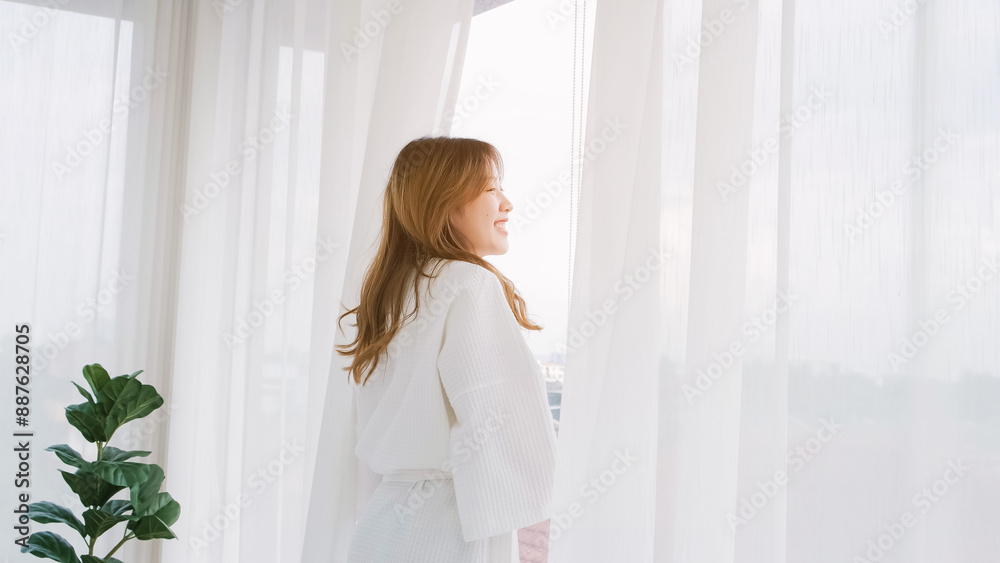 Woman wearing bathrobe opens curtains in bedroom and see around the outdoors. Welcome new sunny day, happy positive young woman wakes up and looks in the window at home or hotel feeling optimistic. Woman wearing bathrobe opens curtains in bedroom and see around the outdoors. Welcome new sunny day, happy positive young woman wakes up and looks in the window at home or hotel feeling optimistic.