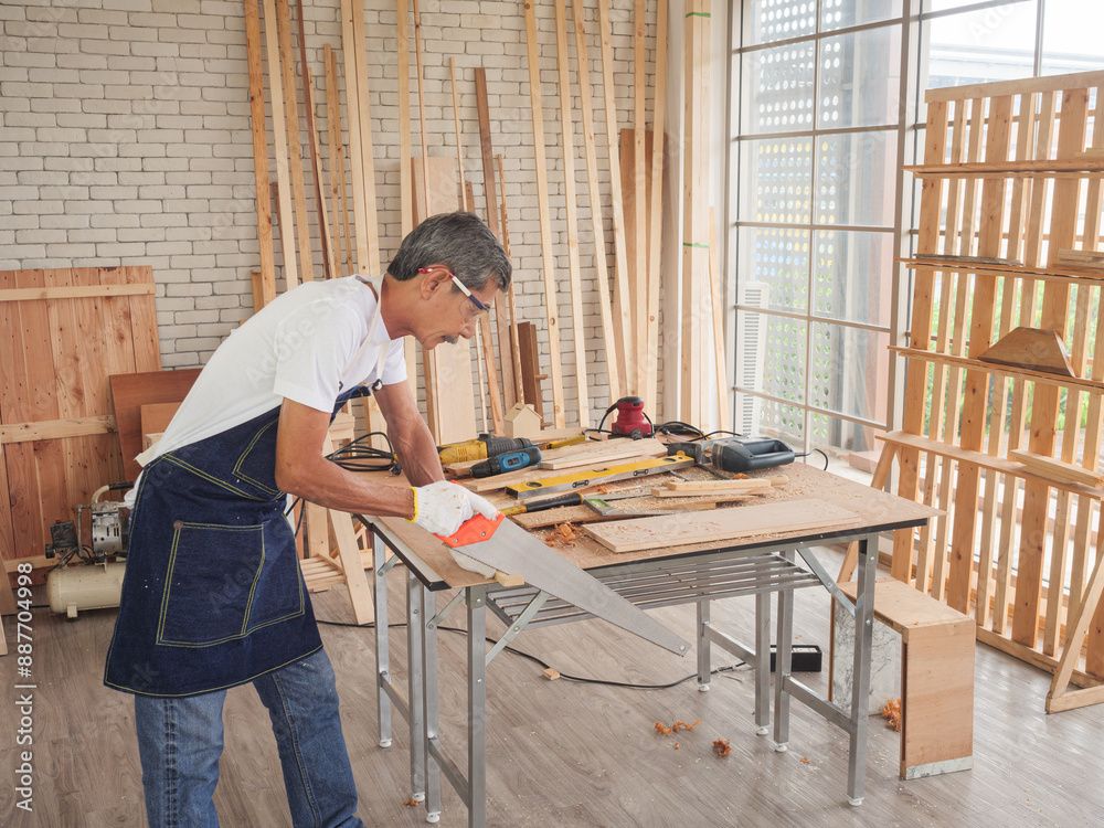 The older carpenter working on woodcraft using a hand saw for handcrafting at the workshop to produce wooden furniture. The older carpenter working on woodcraft using a hand saw for handcrafting at the workshop to produce wooden furniture.