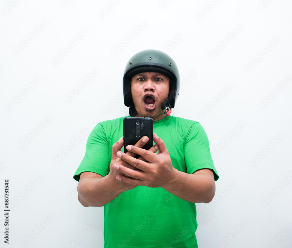 Asian man wearing a helmet and green shirt showing a surprised gesture or expression while holding a mobile phone Asian man wearing a helmet and green shirt showing a surprised gesture or expression while holding a mobile phone