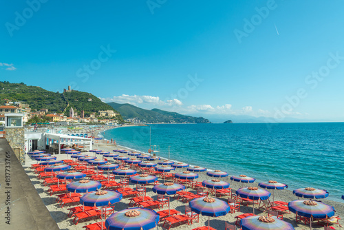 The coastline of Noli, Liguria, Italy, with beach umbrellas in several rows on the sand. Turquoise waters of mediterranean sea on the right. Green hills on the background.