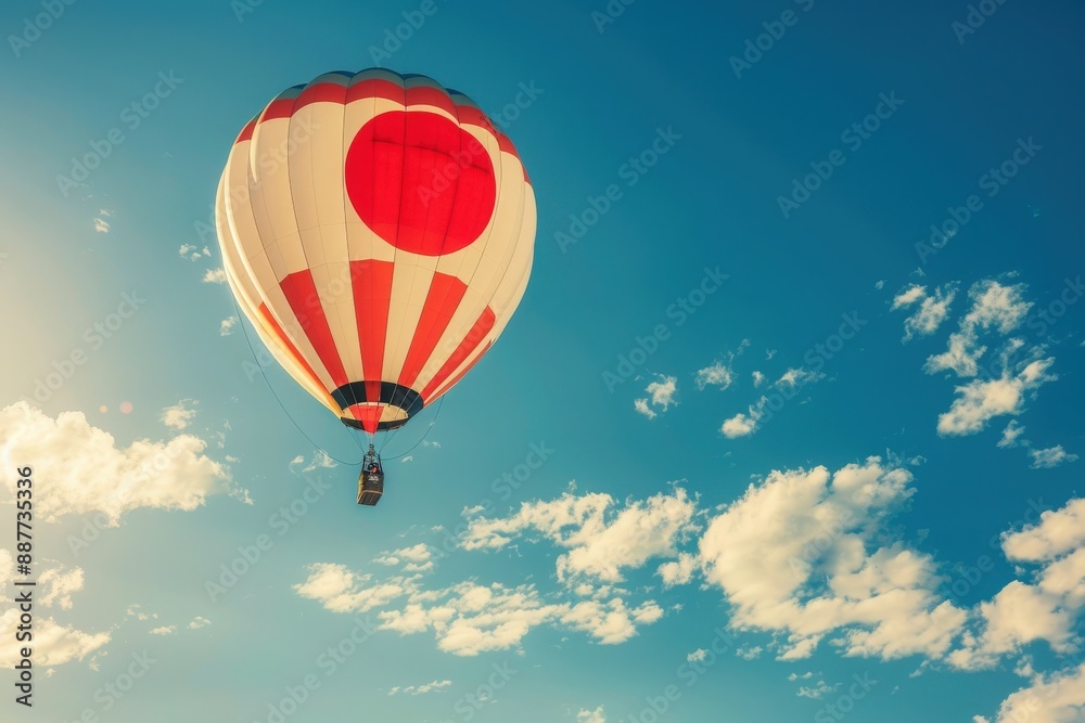 Fototapeta premium A hot air balloon adorned with the Japanese flag ascends against a backdrop of fluffy white clouds and a bright blue sky.