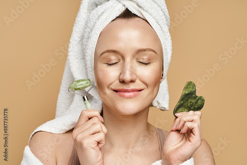 A serene woman with a towel on her head holds a face roller and a, showcasing her natural beauty routine.